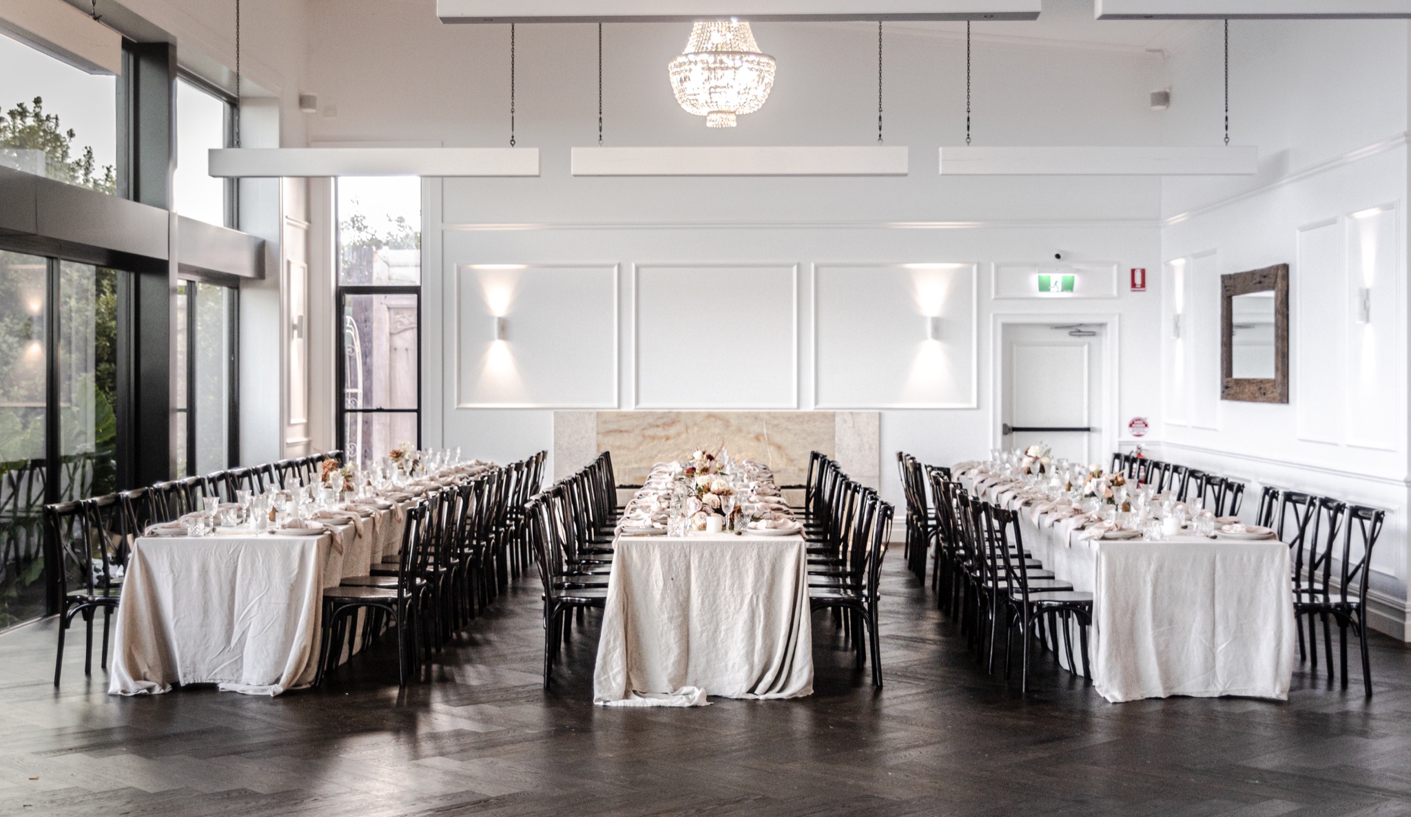The Tides Grand Hall set with long reception tables, black bentwood chairs and chandeliers beside tall windows.